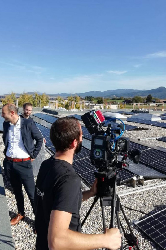 Während einer Filmproduktion bedient ein Kameramann eine Stativ-Kamera auf einem Flachdach, das mit Photovoltaik-Panels bedeckt ist.